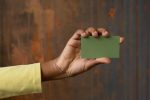 Anonymous African American person showing green blank business card with copy space in hand while standing near wooden wall in studio