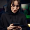 A young woman in a black hoodie uses her smartphone indoors, surrounded by tech gear.