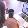 A woman enjoys coffee while browsing the web on a laptop at a home office desk.