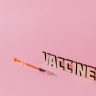 Syringe and 'Vaccine' text on pink background, conceptualizing healthcare and immunization.