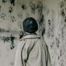 A person stands in a haunting room filled with black handprints, evoking a sense of mystery and fear.