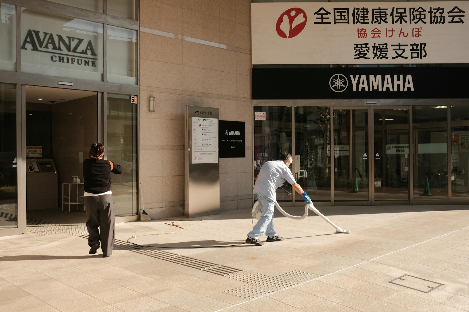 Two people clean the pavement outside a Yamaha building in Japan. Urban maintenance scene.