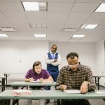 A teacher observes students taking a test in a brightly lit classroom environment.