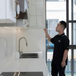 A man in black examines a kitchen cabinet inside a modern apartment with large windows.