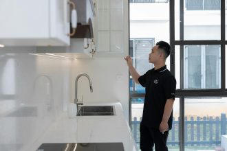 A man in black examines a kitchen cabinet inside a modern apartment with large windows.