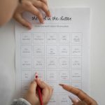 Hands using a red pencil to mark a declutter challenge sheet indoors.