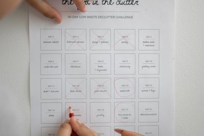 Hands using a red pencil to mark a declutter challenge sheet indoors.