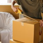 Crop female wearing casual clothes standing while packing carton boxes with scotch tape in modern apartment during preparing for relocation