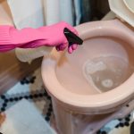A person uses pink gloves and a sprayer to clean a pink toilet, emphasizing cleanliness.