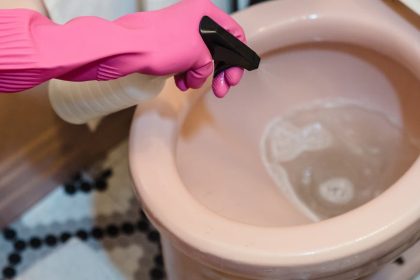 A person uses pink gloves and a sprayer to clean a pink toilet, emphasizing cleanliness.