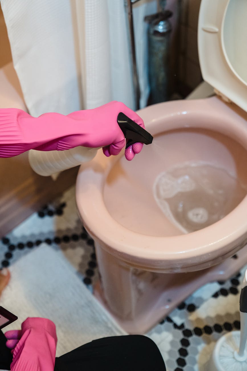 A person uses pink gloves and a sprayer to clean a pink toilet, emphasizing cleanliness.
