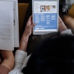 Overhead view of a person reading news on a digital tablet while holding an open book at home.